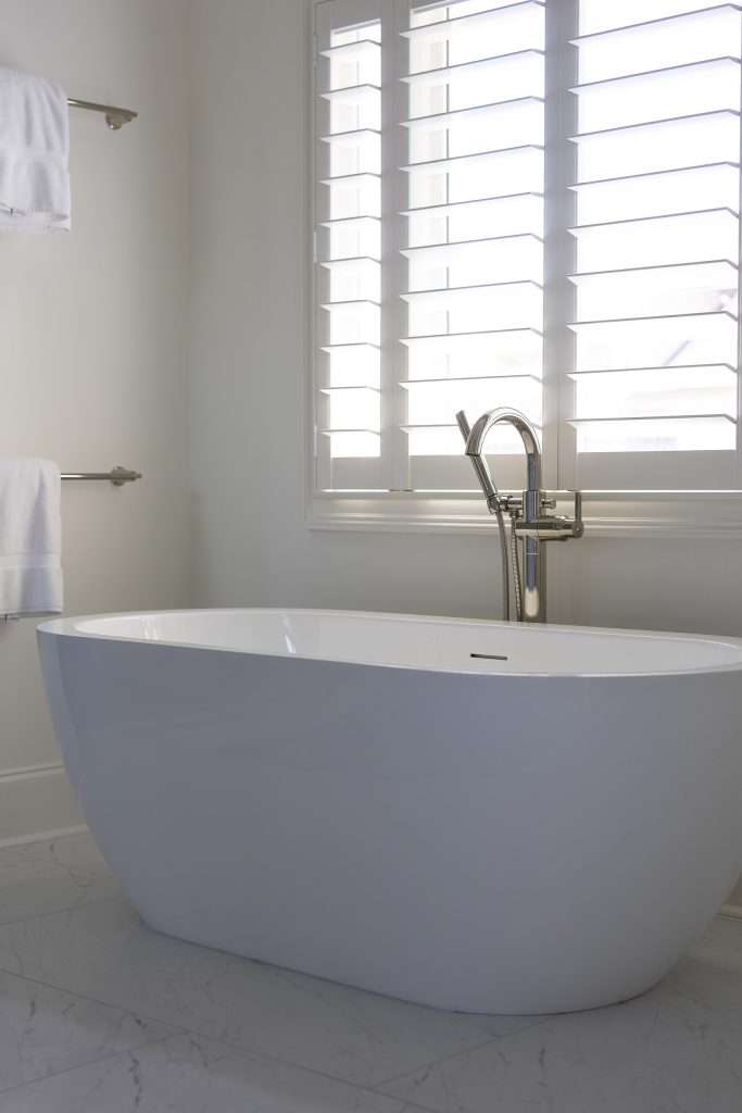 Freestanding white soaking tub beneath a shuttered window with a polished-nickel floor-mounted filler.