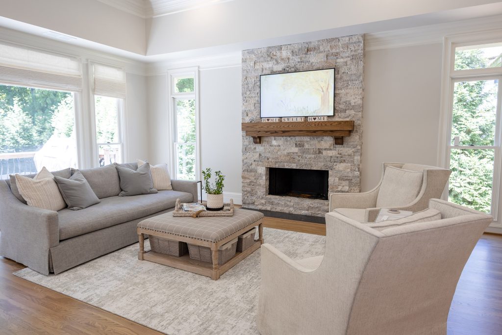 Bright living room with a stacked-stone fireplace, gray sofa, two cream swivel chairs, and abundant windows.