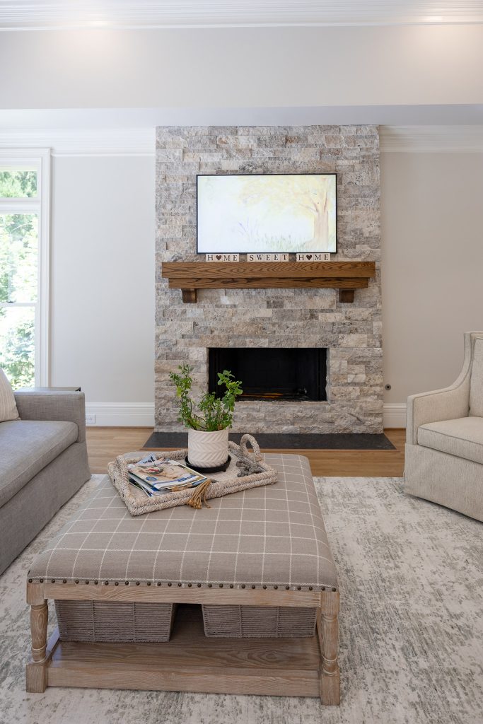 Living room with a stacked-stone fireplace, stained-oak mantel, gray sofa, and a plaid upholstered ottoman.