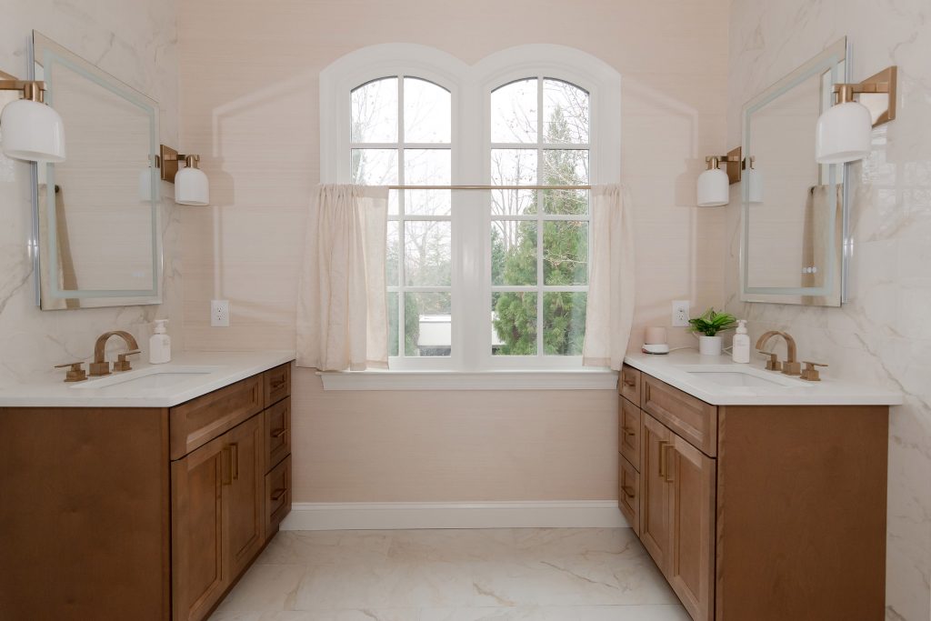 Double-vanity master bath with stained wood cabinets, white quartz tops, gold fixtures, and a central arched window.