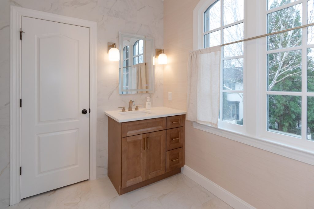 Light bath with a stained wood shaker vanity, quartz top, marble-look tile wall, and an arched window with cafe curtains.