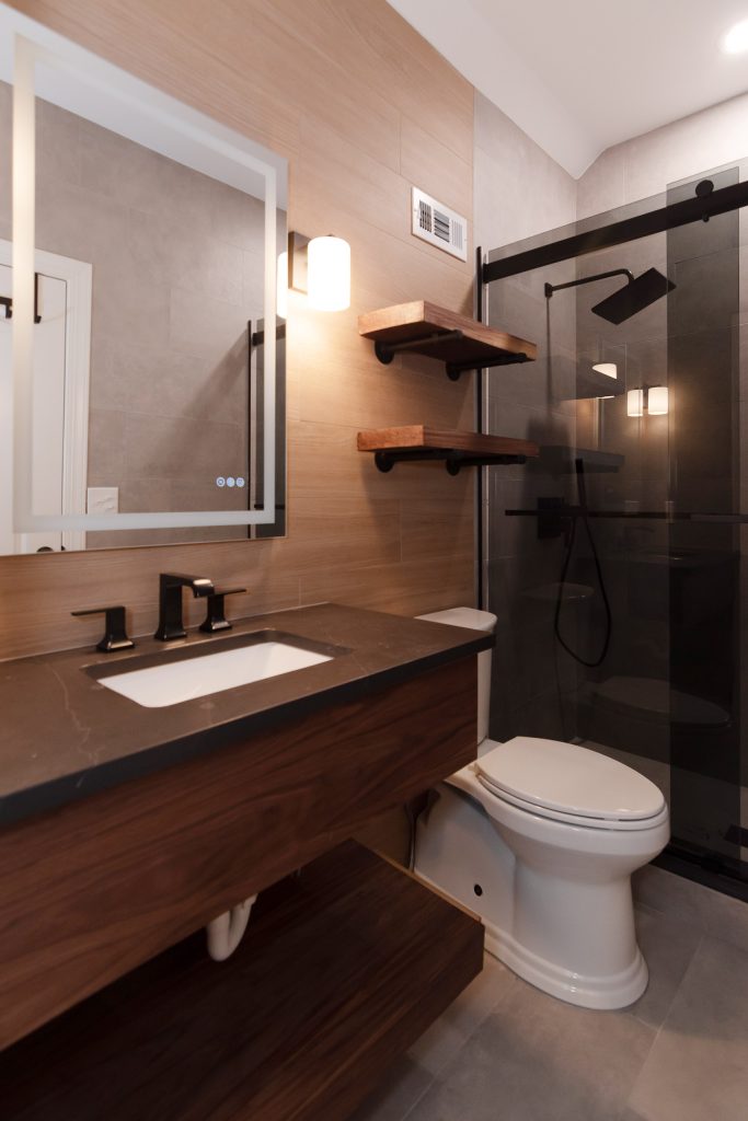 Modern bath with a floating walnut vanity, dark quartz counter, black faucet, live-edge shelves, and a smoked-glass shower.