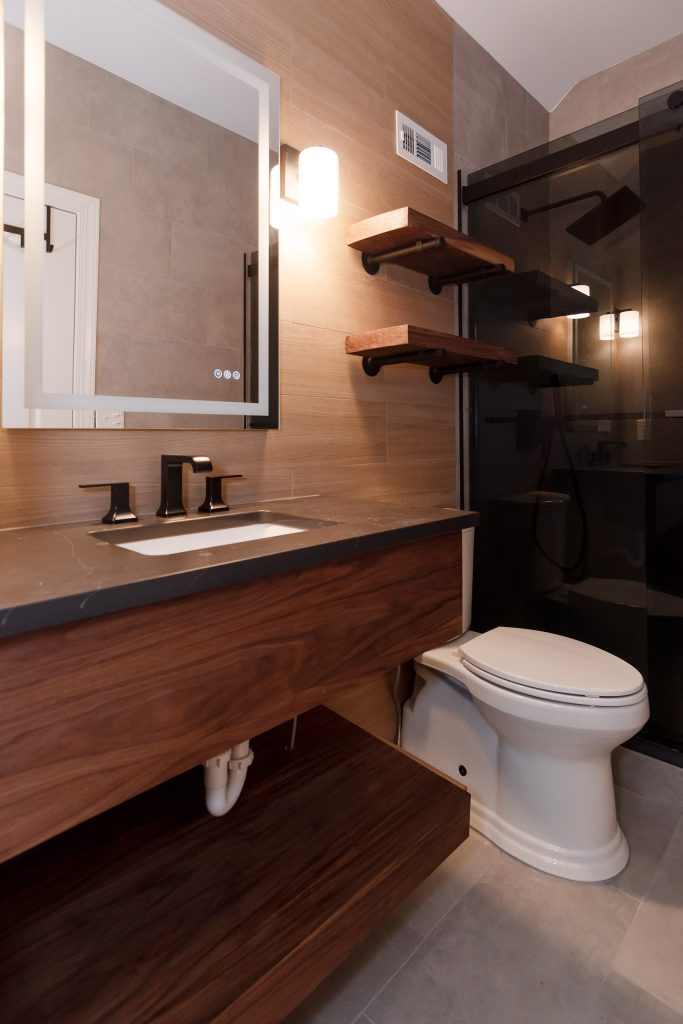 Modern bath with a floating walnut vanity, dark quartz counter, black faucet, live-edge shelves, and a dark-glass shower.