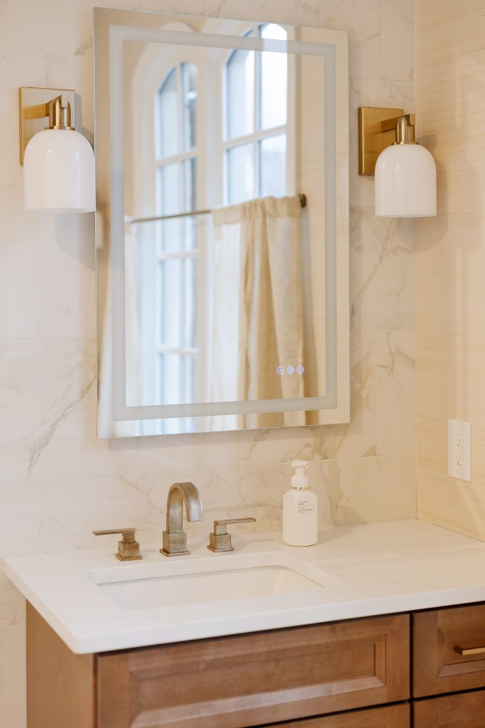 Vanity close-up with a stained wood cabinet, white quartz counter, brushed fixtures, gold sconces, and an LED mirror.