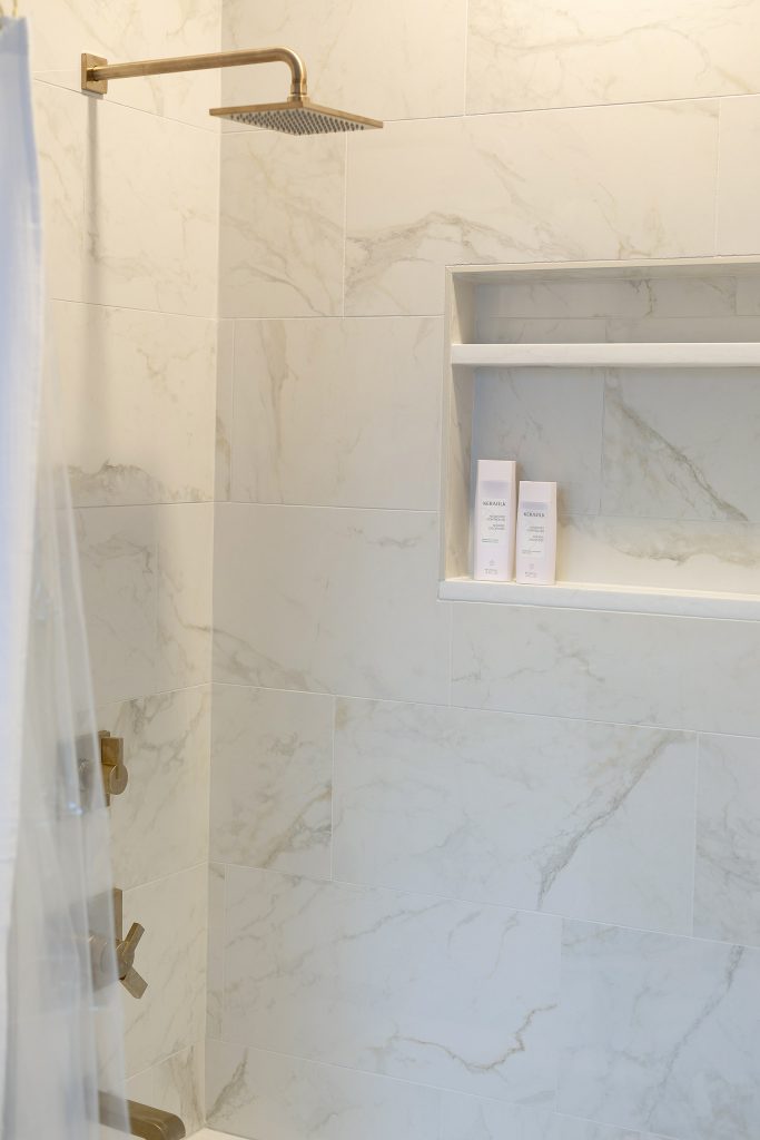 Marble-look shower wall with a brushed-gold rainfall head, cross-handle valve, and a recessed niche holding shampoo.