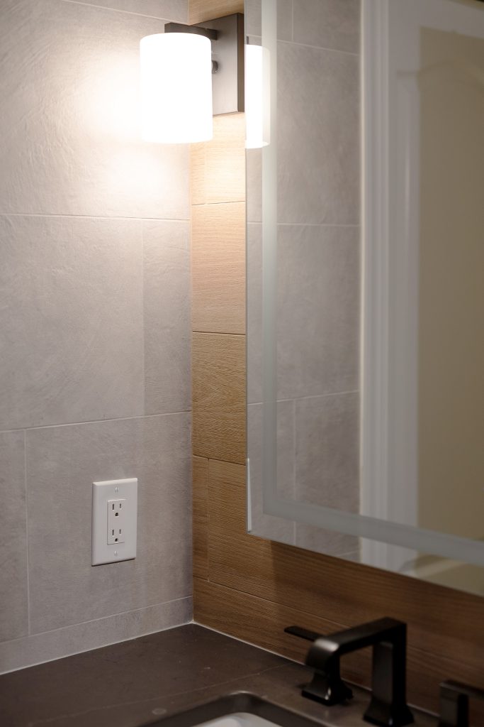 Detail of a modern vanity corner with gray tile, a wood-look tile band, a white sconce, and a black faucet on dark quartz.