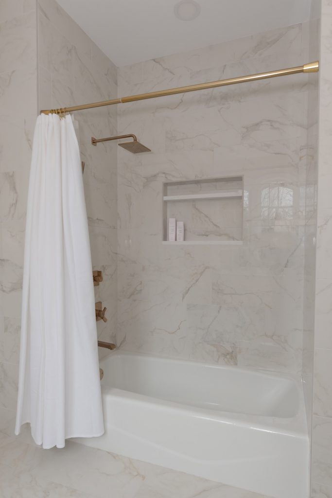 Marble-look tub-shower with a brushed-gold rainfall head, recessed niche, and a white curtain on a gold rod.