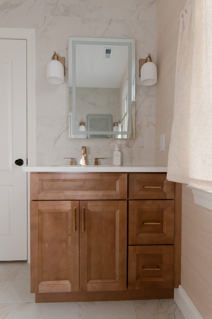 Stained wood vanity with white quartz top, gold-trimmed mirror, white sconces, and a marble-look tile backsplash.