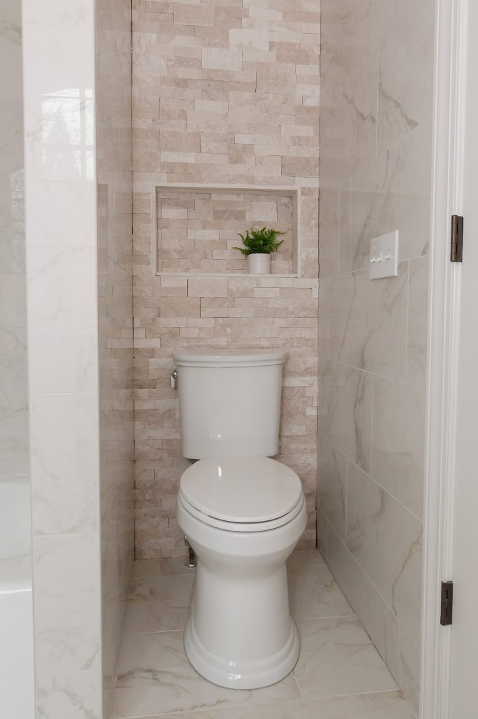Private toilet alcove with a stacked stone feature wall, framed niche and fern, flanked by marble-look tile.
