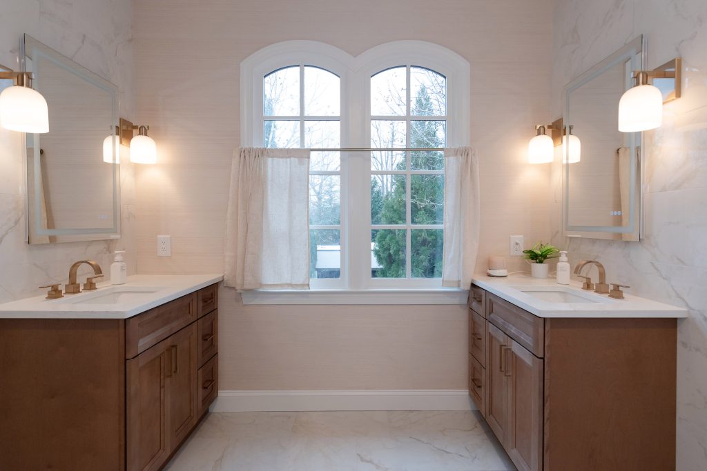 Double-vanity bath with two stained wood cabinets, white quartz tops, gold fixtures, and an arched window between mirrors.