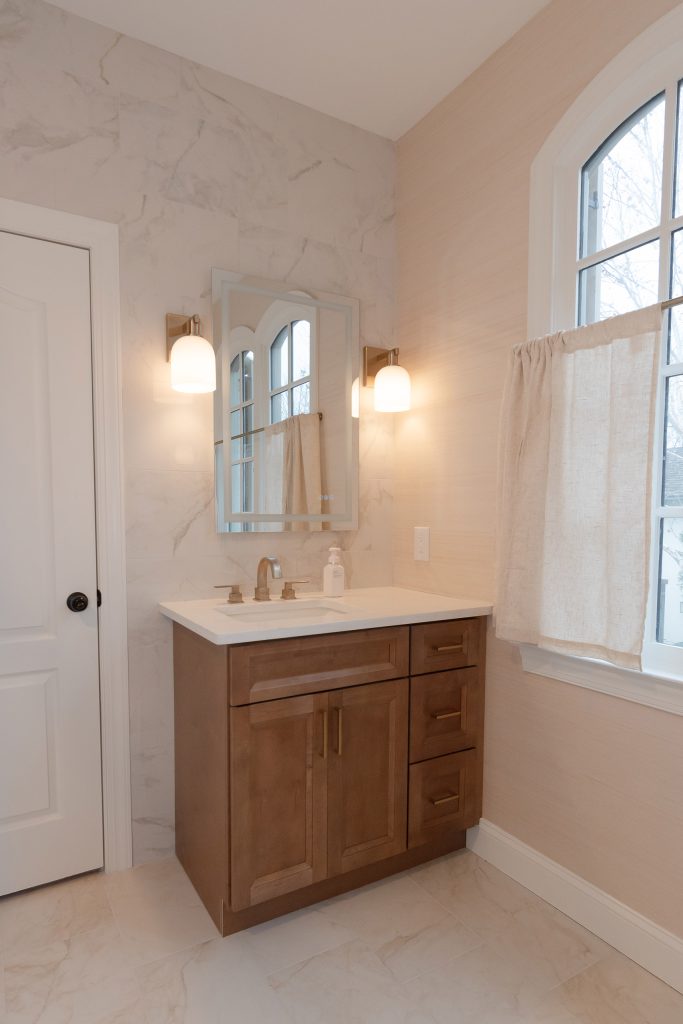 Stained wood vanity with white quartz counter, LED-lit mirror, gold sconces, and an arched window in a bright bath.