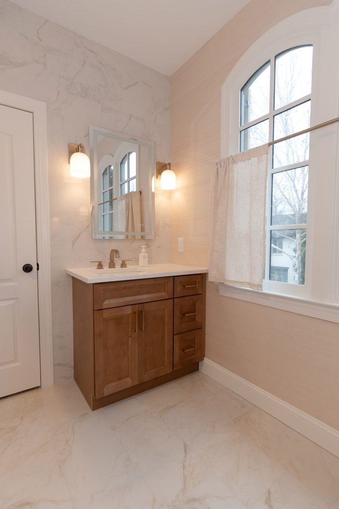Traditional vanity with stained wood cabinets, a white quartz top, marble-look wall, and an arched window with linen curtains.