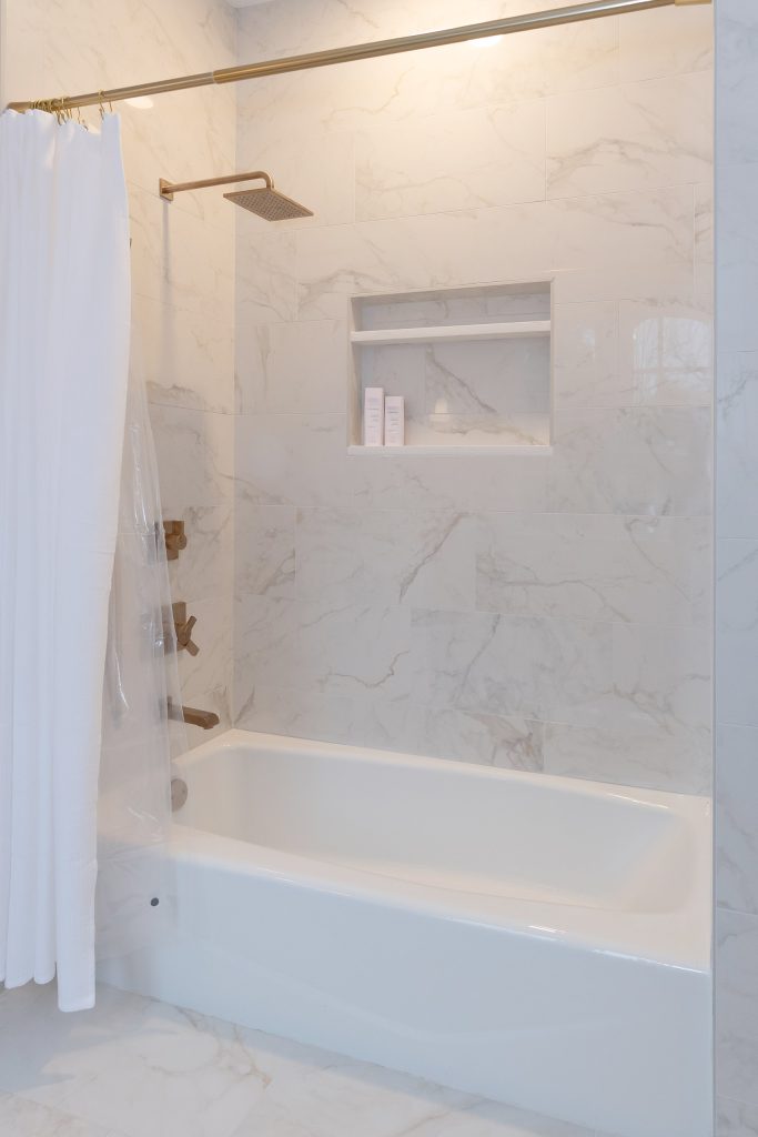 Marble-look tub-shower with a brushed-gold rainfall head, cross handles, a recessed niche, and a white curtain.