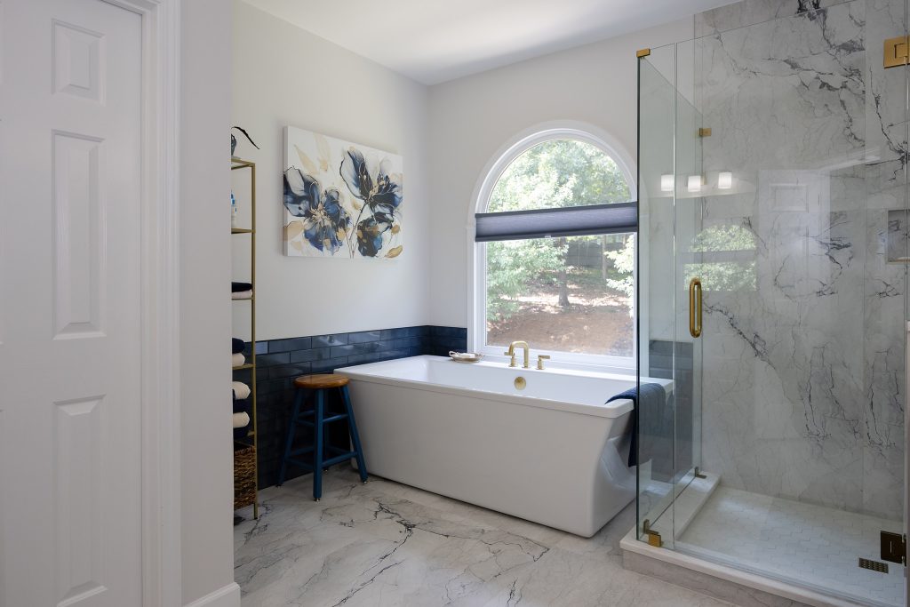 Master bath entry view with freestanding tub, floral canvas, gold ladder shelf, and a marble-look glass shower.