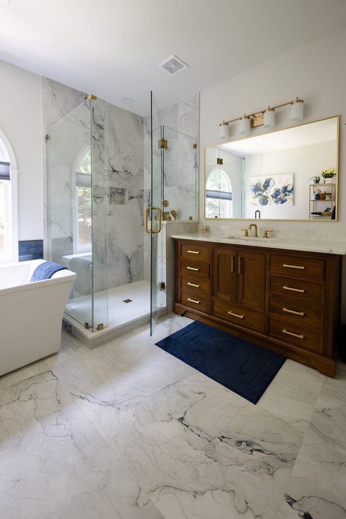 Master bath with freestanding tub, marble-look glass shower, and walnut vanity under a brushed-gold vanity light.