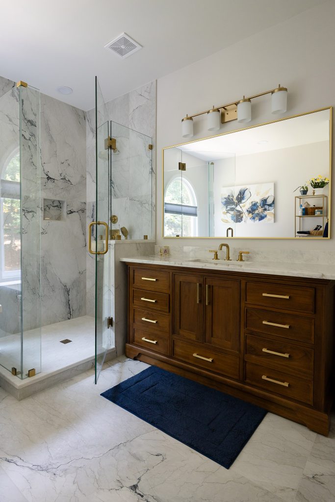 Walnut vanity with brushed-gold pulls and white quartz counter beside a marble-look corner shower.