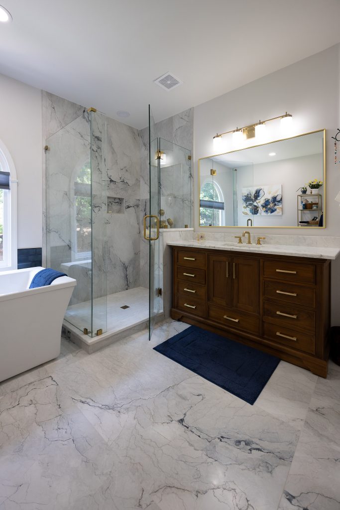 Transitional master bath with corner glass shower, walnut vanity, freestanding tub, and marble-look tile floors.