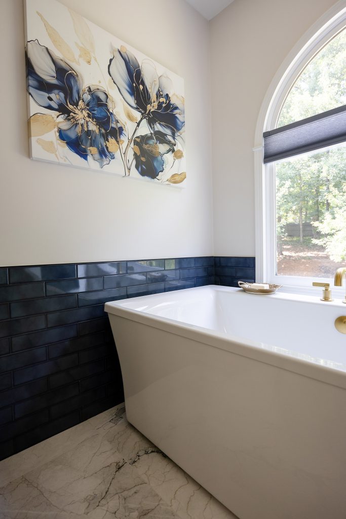 White freestanding tub against navy glossy subway tile with a blue-and-gold floral canvas and an arched window.
