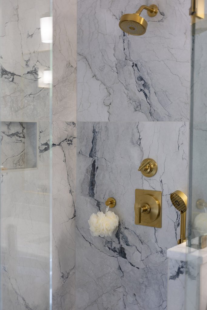 Marble-look shower wall with a gold showerhead, gold sconce, square gold valve, and a hand-held sprayer.