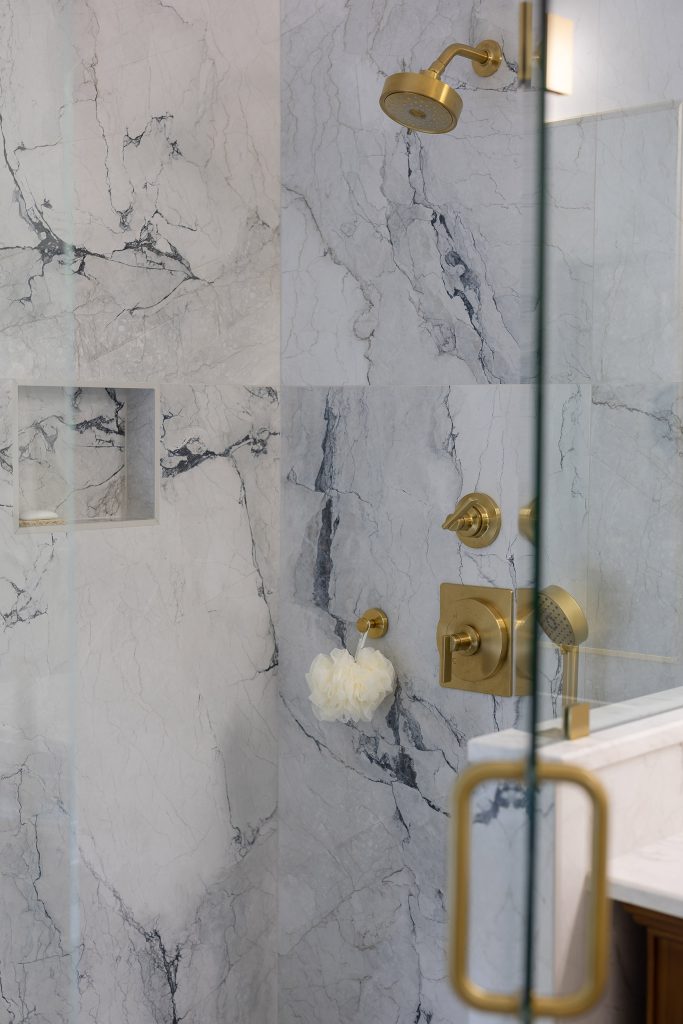 Shower wall of white and blue-gray marble-look tile with a gold showerhead, gold controls, and a hand shower.