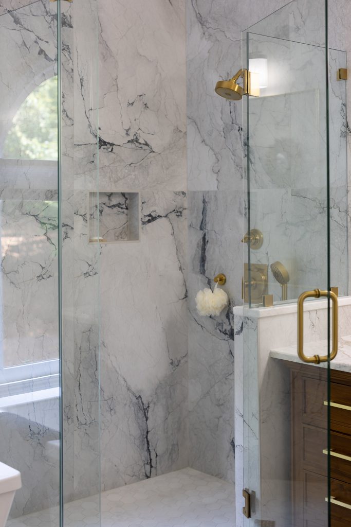 Frameless glass shower with dramatic white and blue-gray marble-look walls, a gold showerhead, and a corner niche.