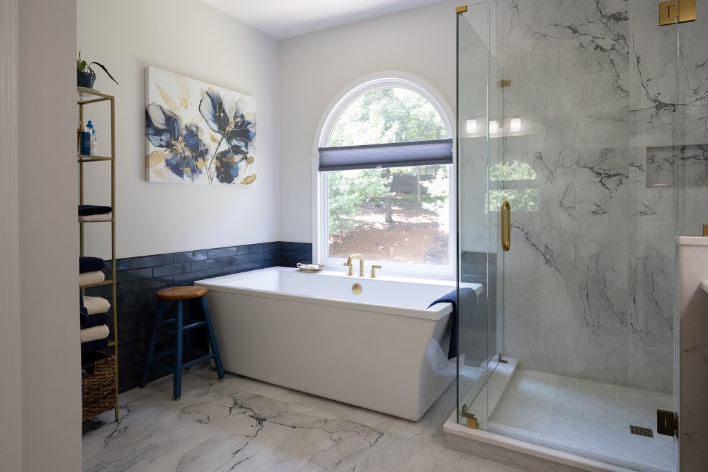 Freestanding tub with a blue stool and gold ladder shelf beside a marble-look glass shower under an arched window.