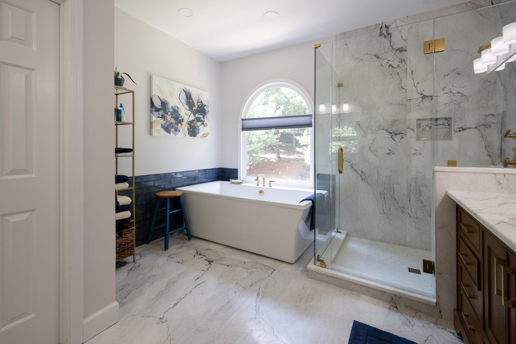 Master bath entry view with freestanding tub, blue stool, gold ladder shelf, and a marble-look glass shower.