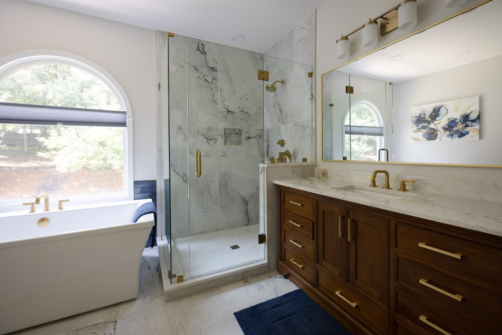 Master bath with freestanding tub, marble-look glass shower, and walnut vanity under a gold-framed mirror.