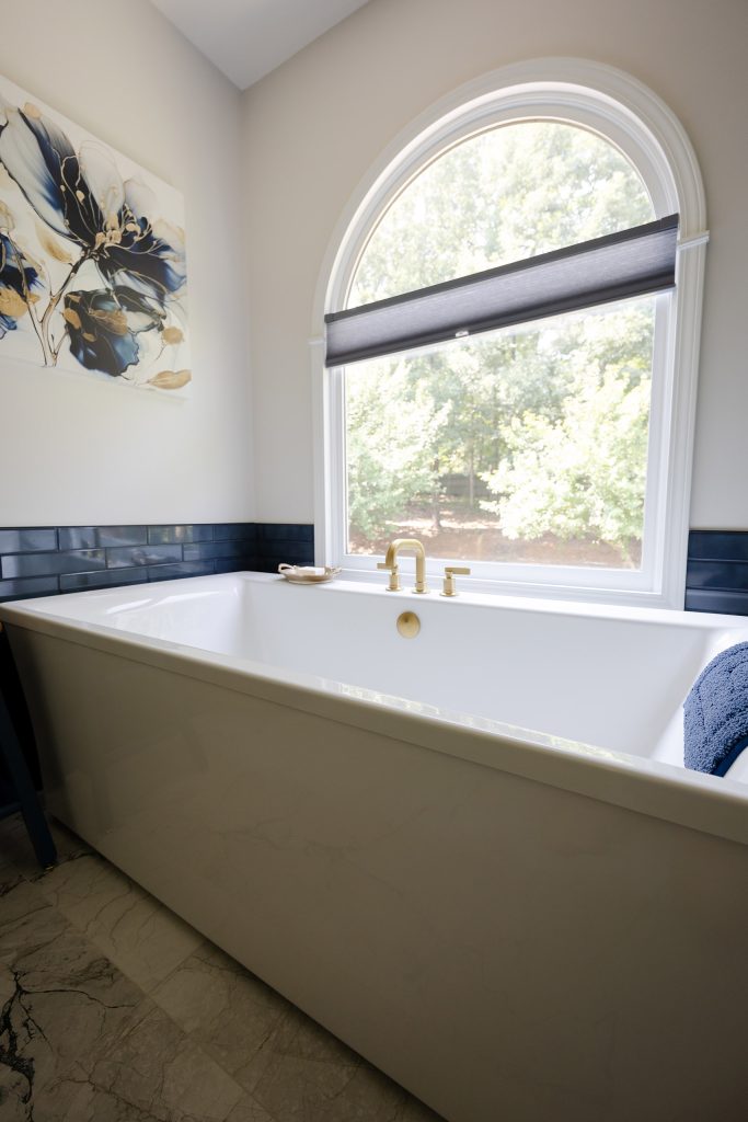 White freestanding tub below an arched window, with a gold deck faucet, navy glossy tile, and a floral canvas.