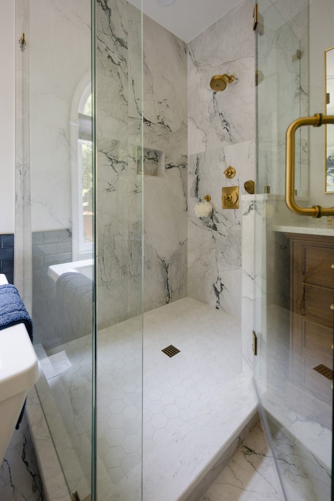Shower interior with hexagon mosaic floor, gold square drain, gold valve, and an arched window in marble-look tile.