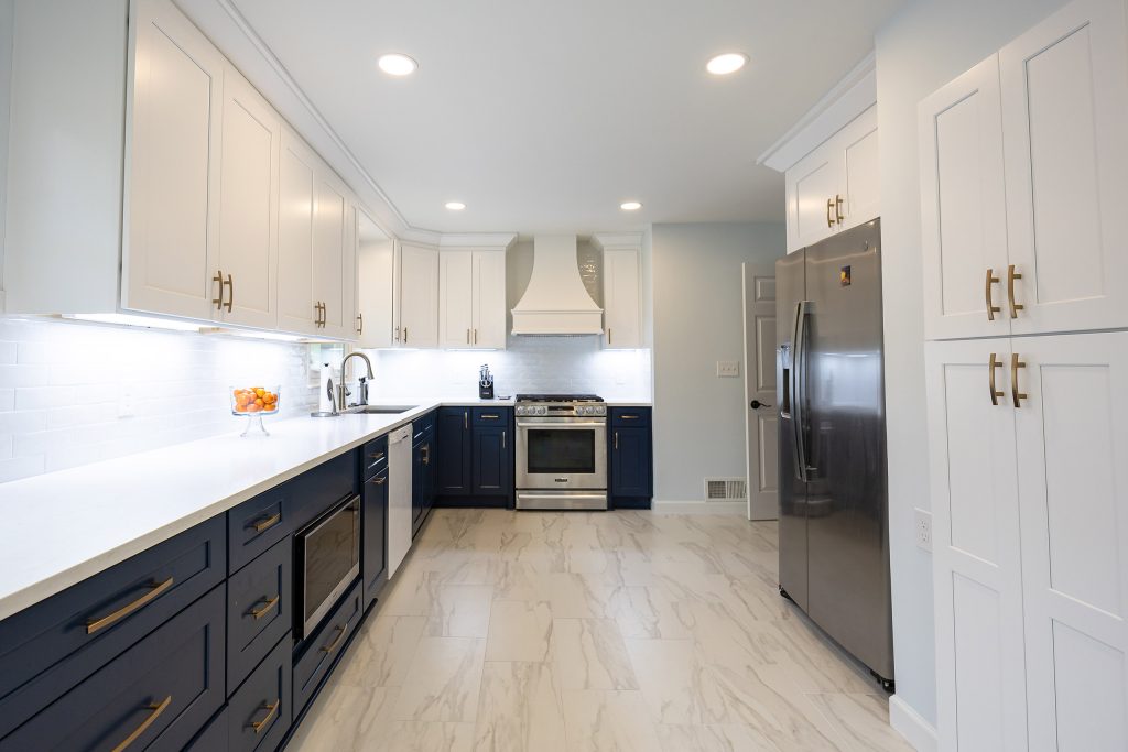 Open two-tone kitchen with navy lowers, white uppers and pantry, a custom white hood, and a stainless refrigerator.