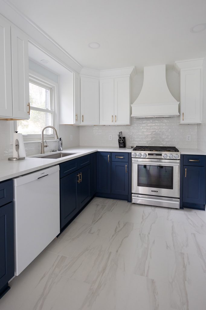 Two-tone kitchen with navy L-shaped base, white uppers, a window over the sink, and a stainless range under a white hood.