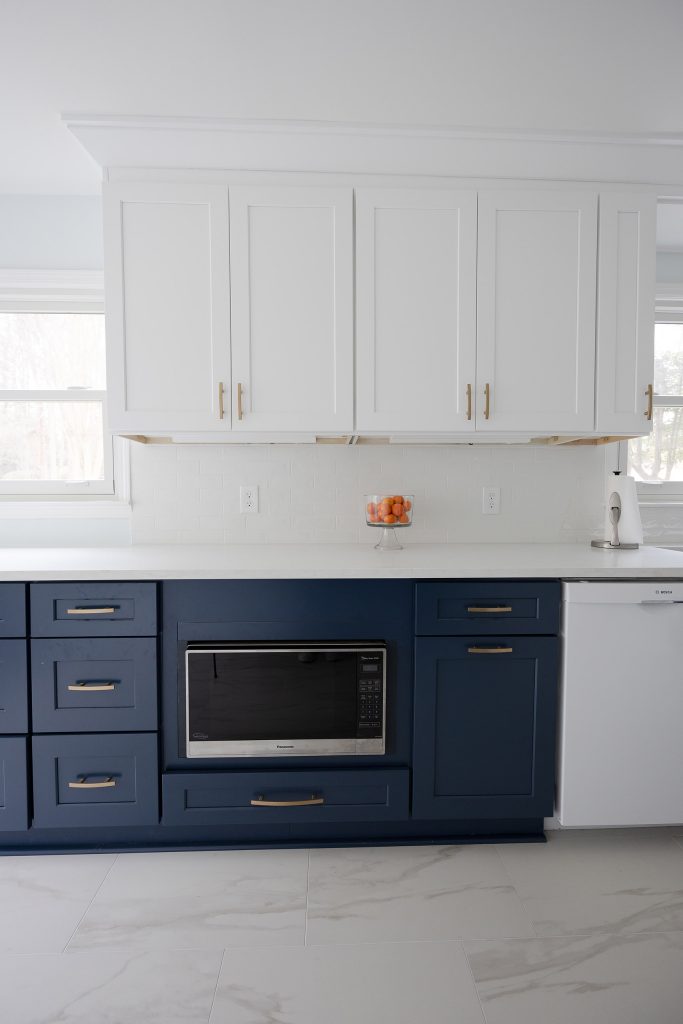 Navy shaker base cabinets with brushed-gold pulls and a built-in microwave, white uppers above, and oranges on white quartz.