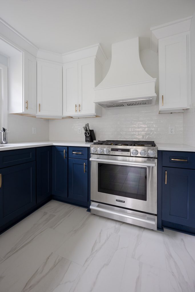 Corner of a two-tone kitchen with a stainless range, custom white hood, navy lowers, and white uppers over marble-look tile.