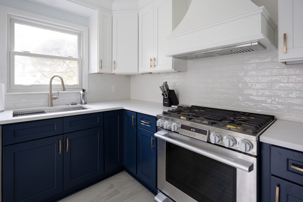 Marietta kitchen with navy blue lower cabinets, white uppers, brushed gold hardware, a white shaker hood, glossy subway tile backsplash, and a stainless gas range.