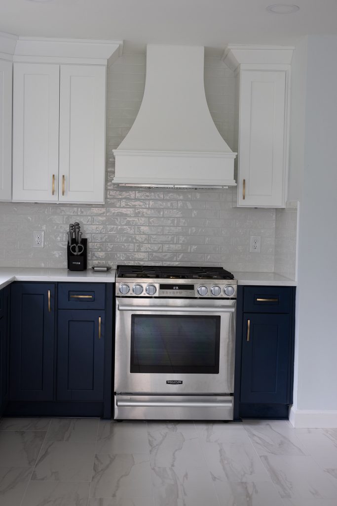 Stainless gas range centered under a custom white hood between navy base cabinets and a glazed subway-tile backsplash.