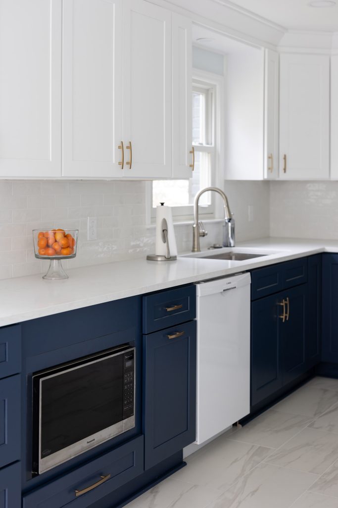 Two-tone cabinetry with white uppers, navy lowers, a built-in microwave, white dishwasher, stainless sink, and oranges on quartz.