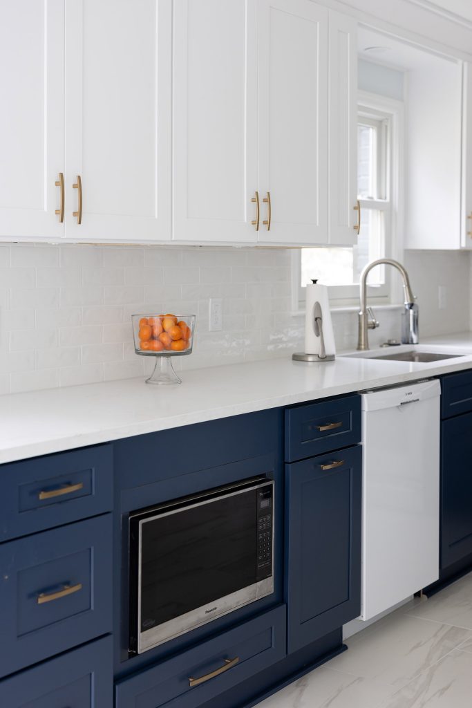 Navy base cabinets with brushed-gold pulls, a built-in microwave, white dishwasher, stainless sink, and a bowl of oranges.