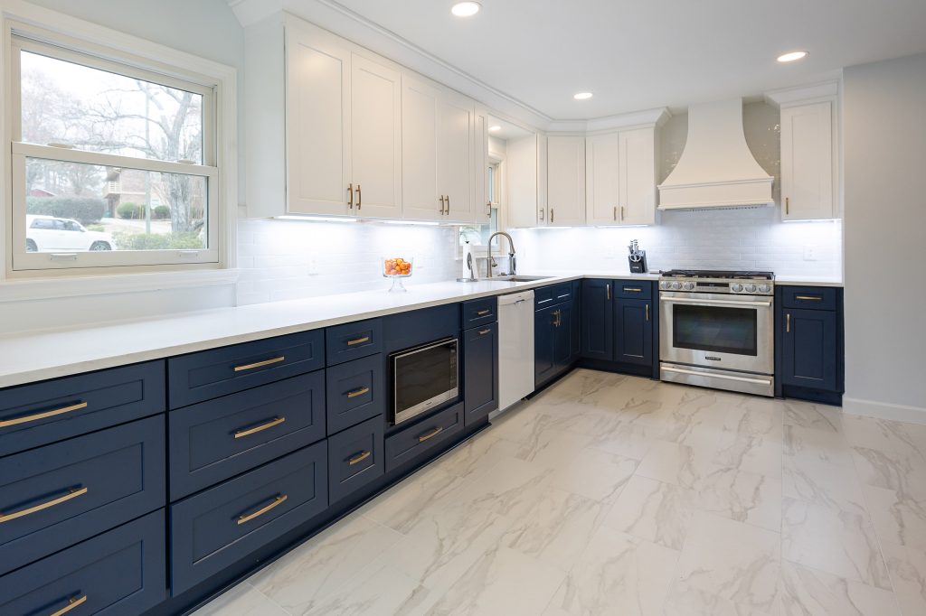 Long counter run of navy shaker drawers with brushed-gold pulls, white uppers, a window, and a stainless range beyond.