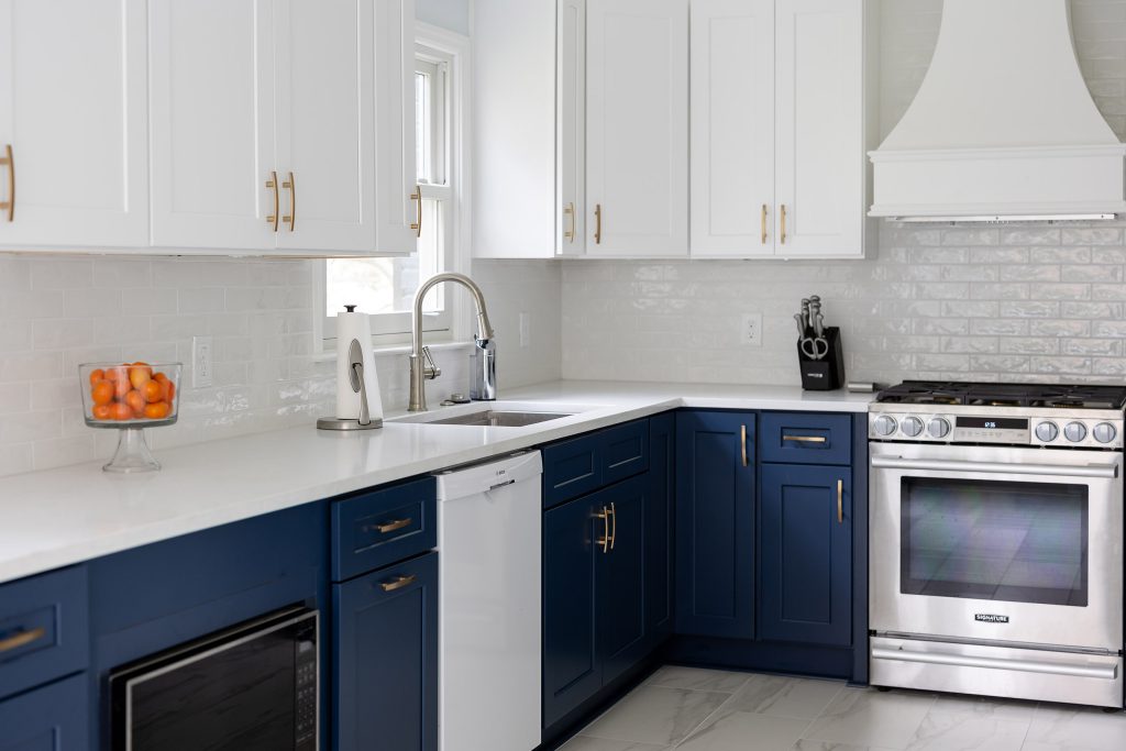 Corner of a two-tone kitchen with a stainless sink, navy cabinets, white uppers, and a stainless range under a white hood.