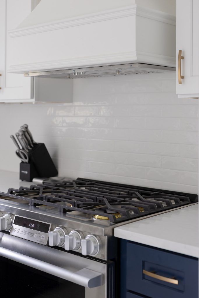 Detail of a stainless gas range's cast-iron grates and burners against a glazed subway backsplash below a white hood.