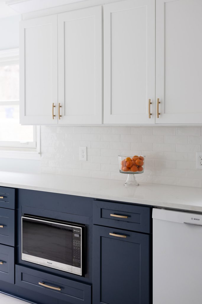 Close-up of white shaker uppers and navy lowers with brushed-gold pulls, a built-in microwave, and a bowl of oranges on quartz.