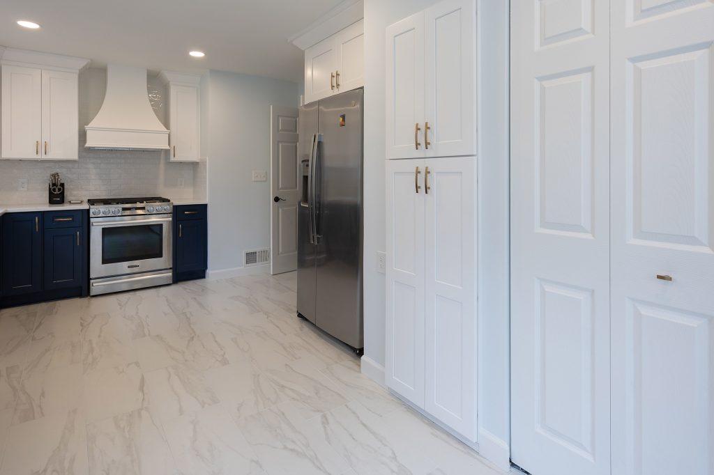 Kitchen floor view showing marble-look tile, tall white pantry cabinets, a stainless refrigerator, and navy cabinets at left.