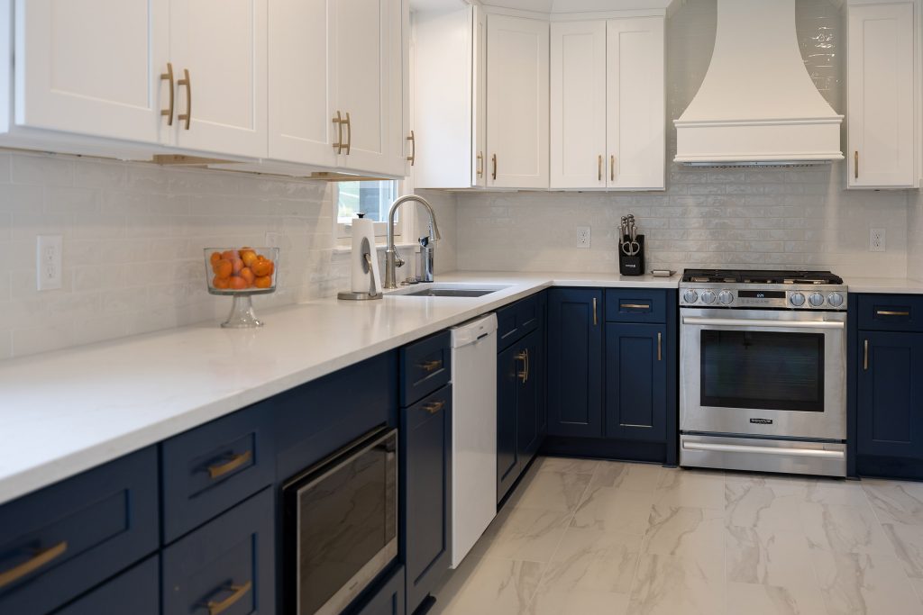 Angled kitchen view with navy base cabinets, white uppers, a bowl of oranges, and a stainless range under a white hood.