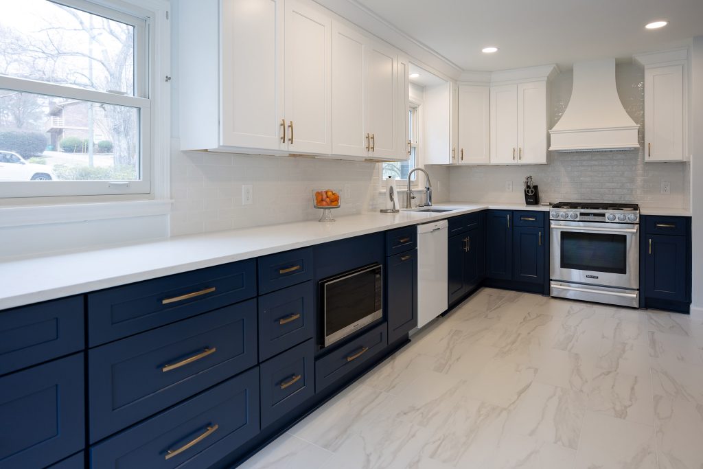 Long run of navy shaker drawers with brushed-gold pulls, white quartz counters, and a built-in microwave below the window.