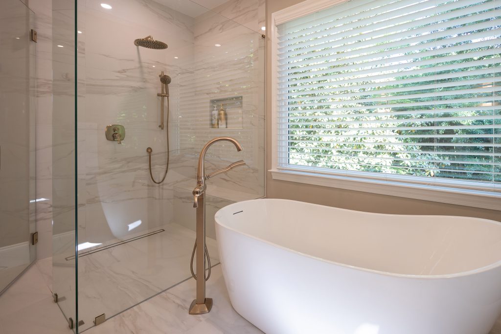 Marble-look walk-in shower with brushed-gold rain head and hand shower beside a white freestanding tub.
