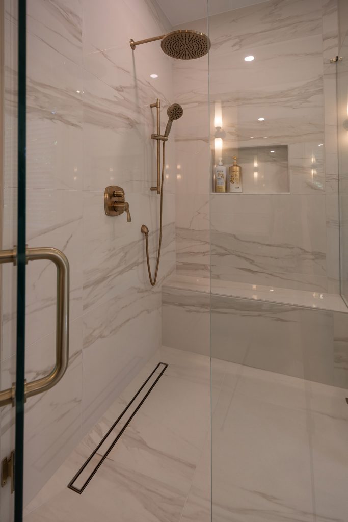 Marble-look shower interior with brushed-gold rain head, hand shower, control, niche, and linear drain.