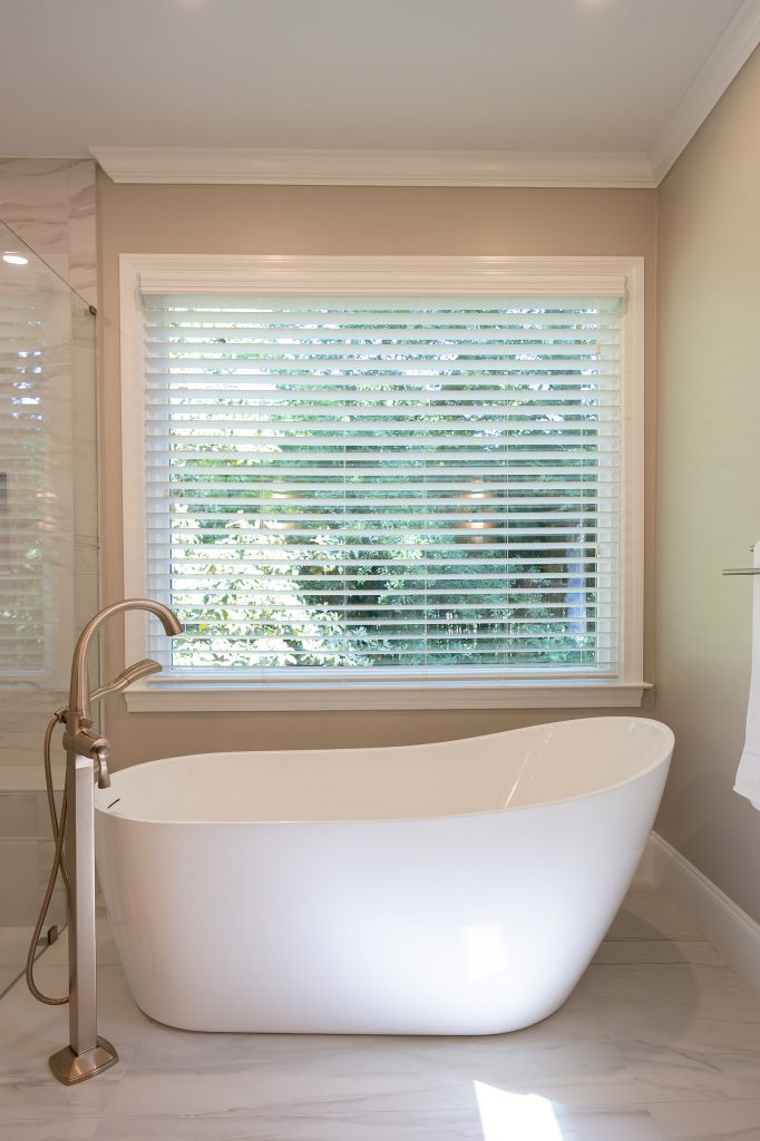 White freestanding slipper tub centered beneath a blinded window with a brushed-nickel floor-mount filler.