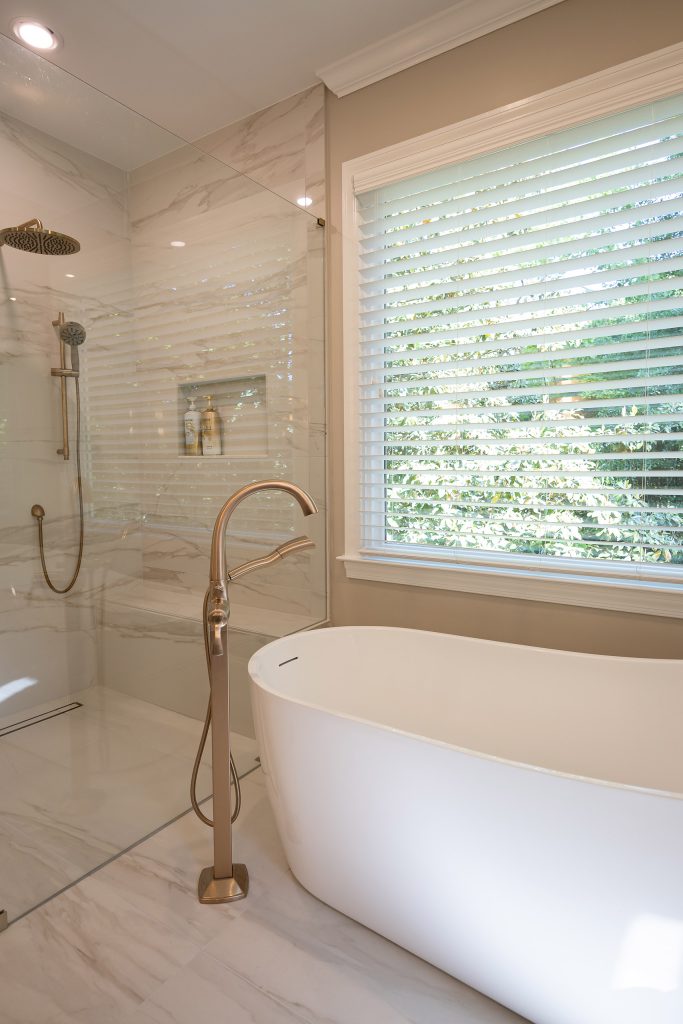 White freestanding tub below a large window beside a marble-look walk-in shower with brushed-gold hardware.