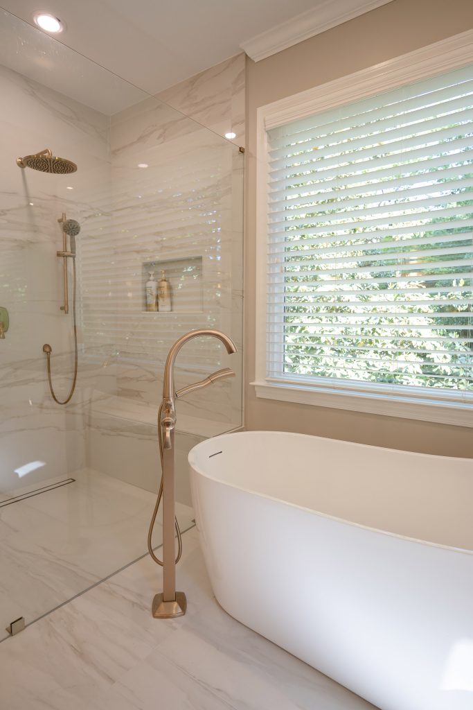 Freestanding soaking tub at a window next to a marble-look walk-in shower with brushed-gold fittings.